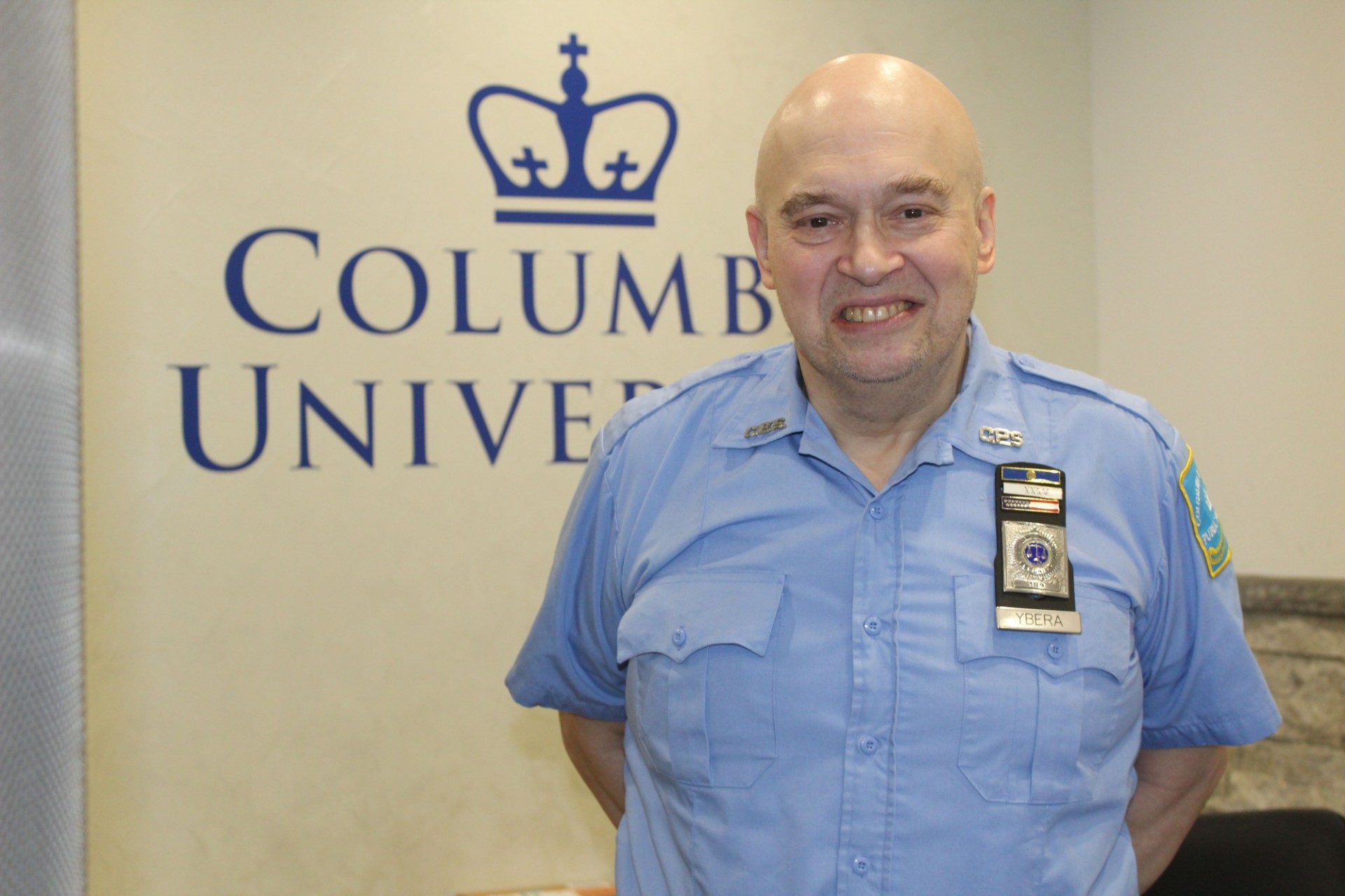 Public Safety Security Officer Benny Ybera poses for a photo in front of white wall with Columbia University logo.