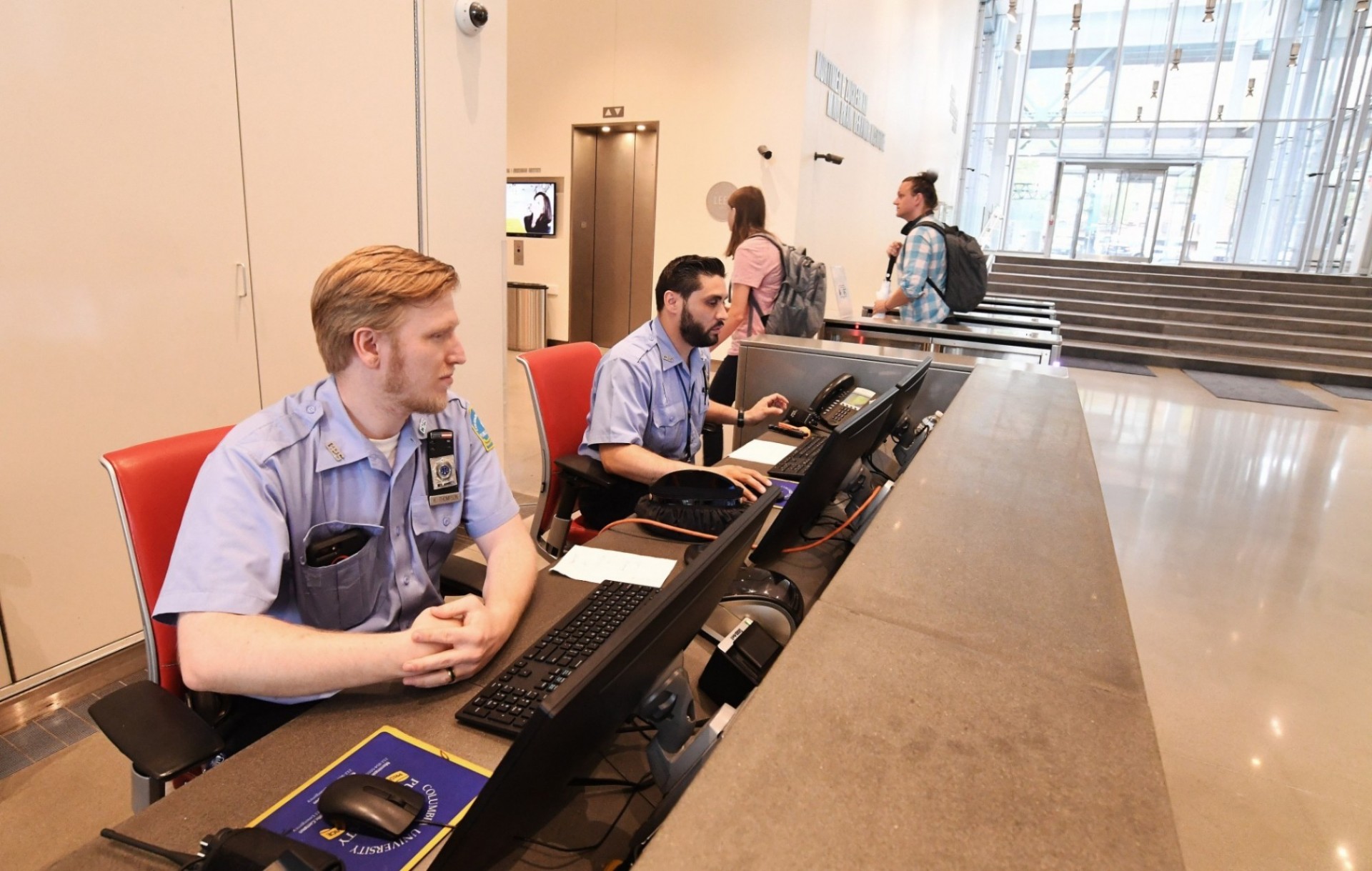 Two officers at the security desk at the Jerome L. Greene Science Center with two people in the background behind the desk entering the turnstiles.