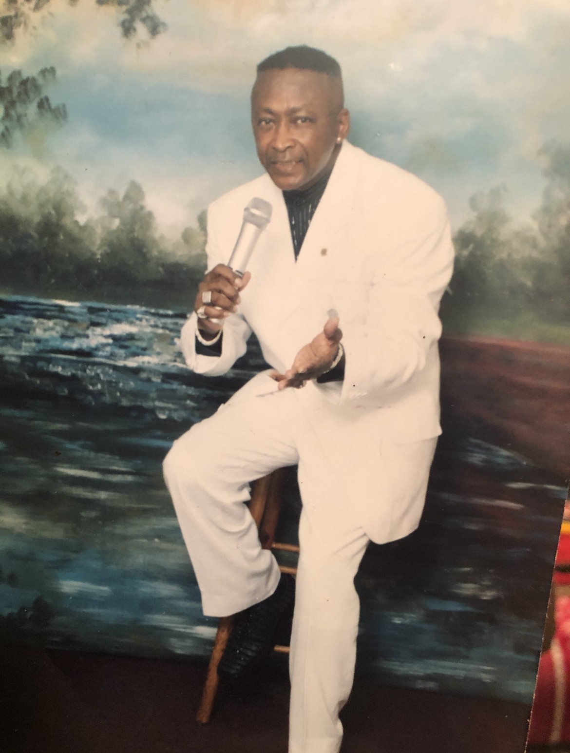 man in a white suit, holding a microphone and sitting on a stool